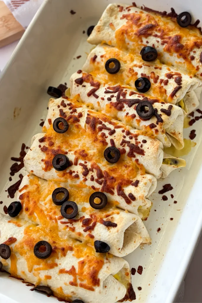 A birds-eye view of breakfast burritos topped with a crispy cheese crust and olives in a baking dish after baking.