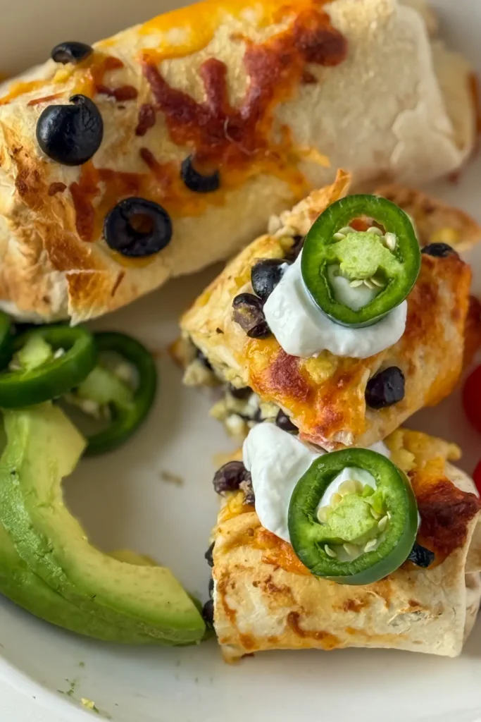 Two crispy burritos, one sliced in half, on a plate with Greek yogurt, avocado and jalapenos.