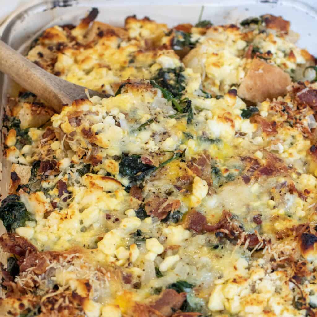 A close-up of bacon, spinach and feta breakfast strata casserole with a wooden spoon in it.