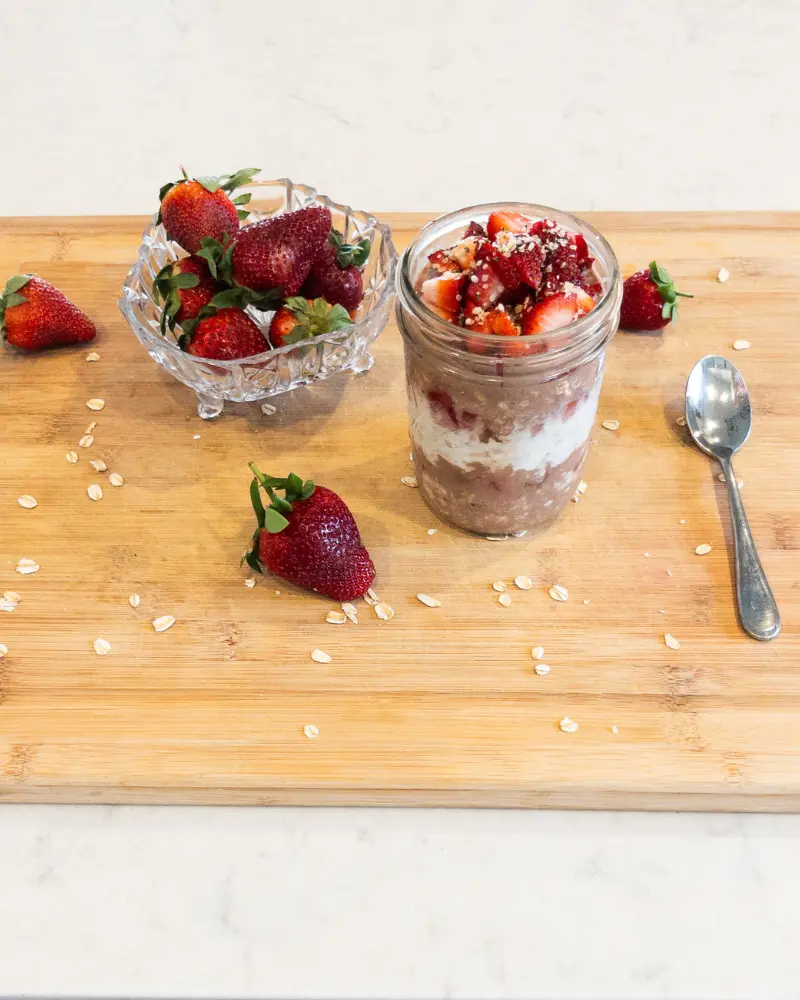A jar of chocolate covered strawberry overnight oats on a wood cutting board next to strawberries and a spoon.