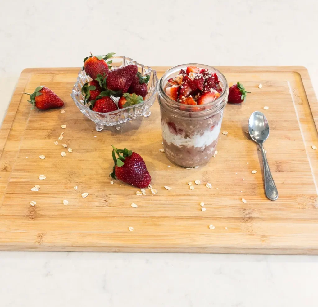 Chocolate covered strawberry overnight oats on a wood cutting board next to a spoon and a bowl of strawberries.