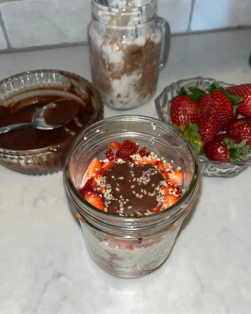 Overnight oats topped with diced strawberries and a healthy dark chocolate date sauce with a bowl of chocolate sauce and a bowl of strawberries behind it on a counter.