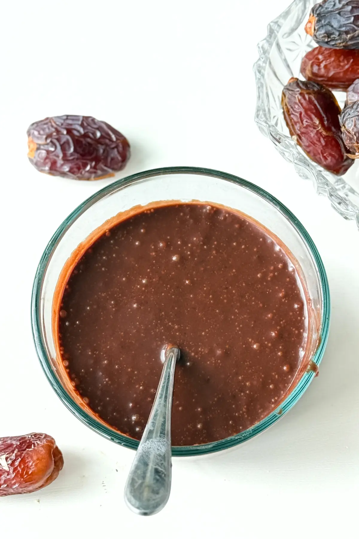 A bowl of dark chocolate date sauce with a spoon in it on a counter next to a bowl of dates.