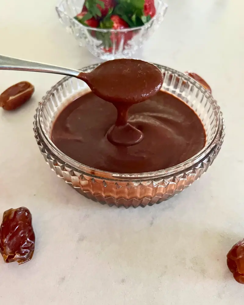 A large spoonful of healthy date chocolate sauce dripping into a bowl of sauce on a counter next to a bowl of strawberries.