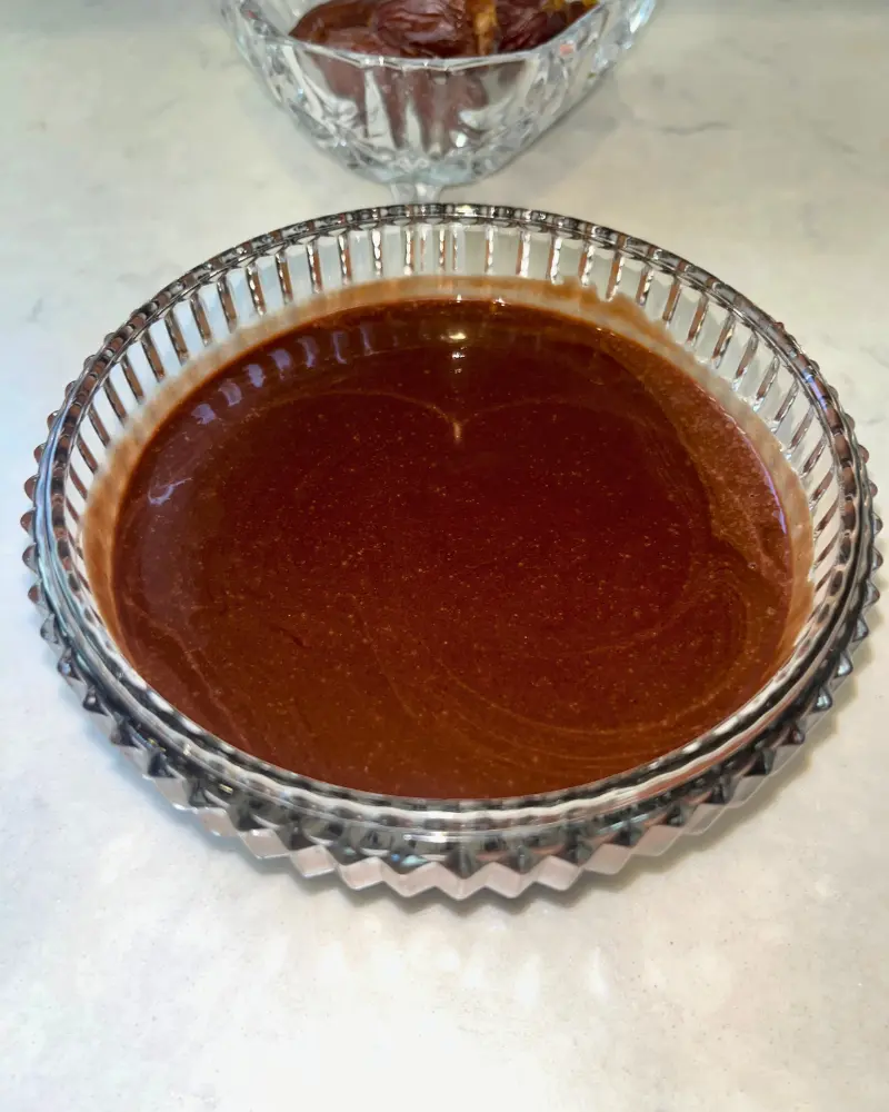 A close-up of a glass bowl of dark chocolate sauce with dates.