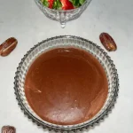 A glass bowl of healthy date chocolate sauce on a counter next to a bowl of strawberries.