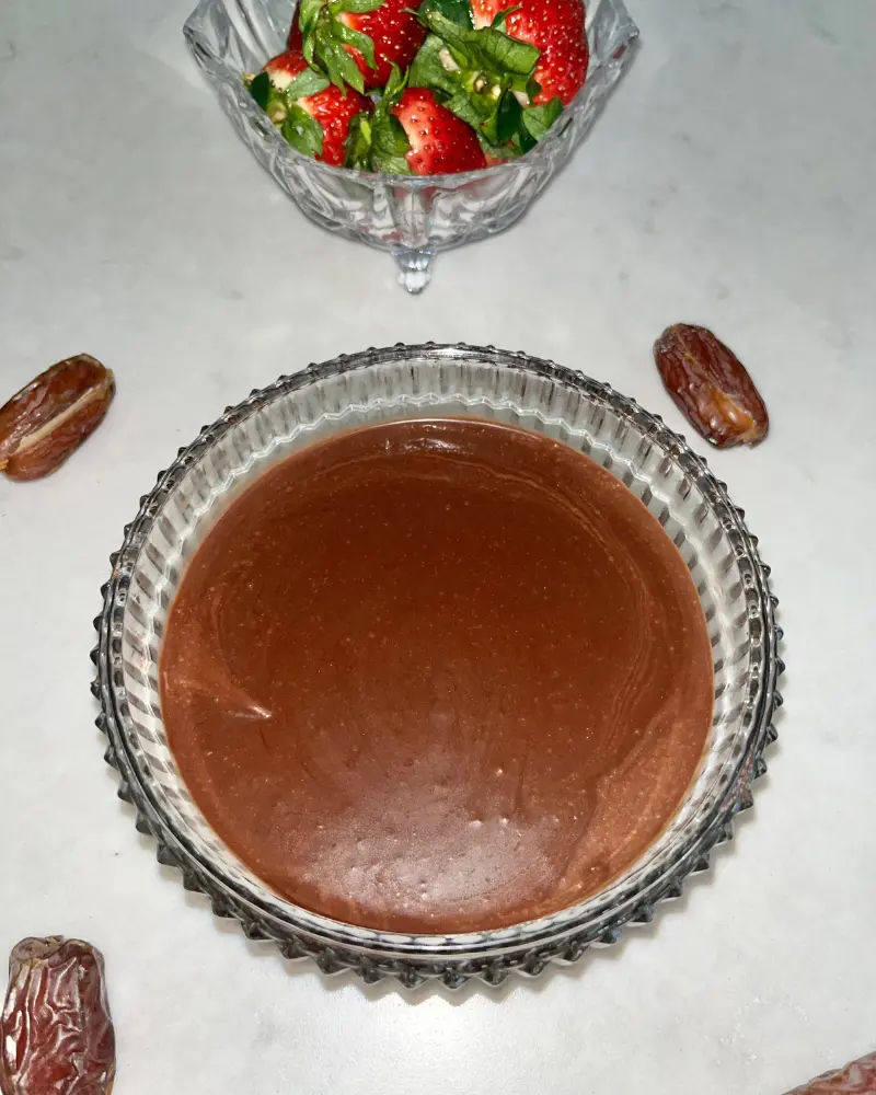 A glass bowl of healthy date chocolate sauce on a counter next to a bowl of strawberries.