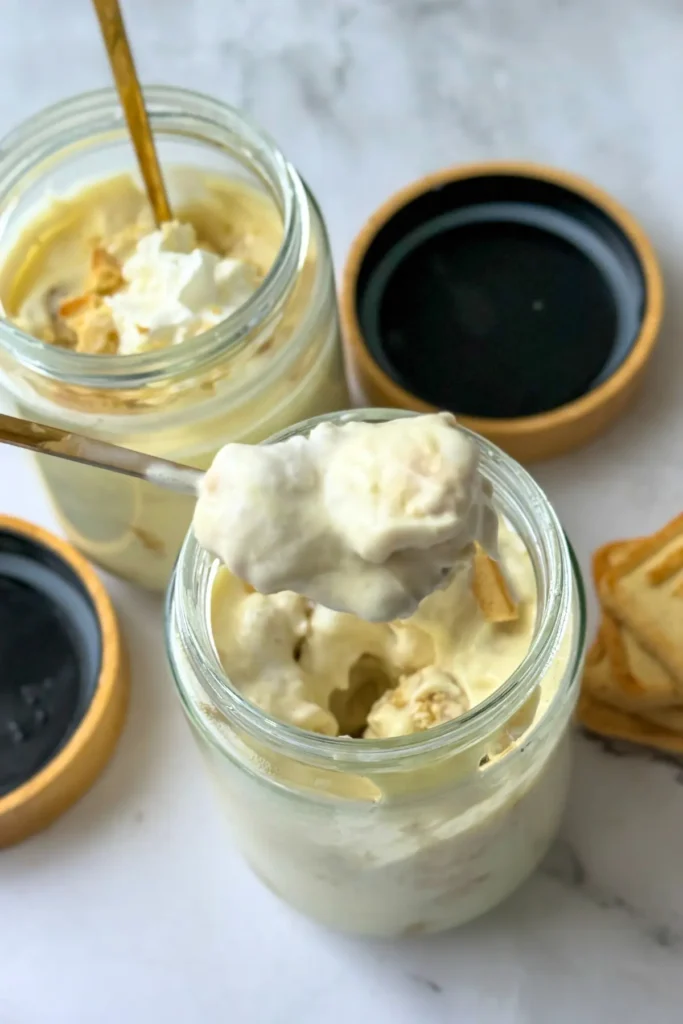 A close-up of a big spoonful of banana pudding over a jar of it on a counter.