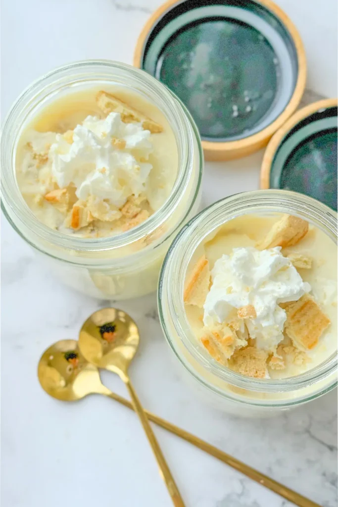 A birds-eye view of two fully assembled Chessmen banana puddings in jars topped with whipped cream and cookies next to spoons on a counter.