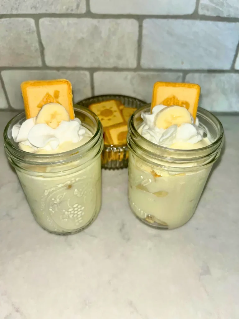 Two jars of high protein banana pudding garnished with a chessmen cookie with a bowl of chessmen cookies in the background.