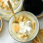 A birds-eye view of two Chessmen butter cookie high protein banana puddings in jars topped with whipped cream with gold spoos on each jar.