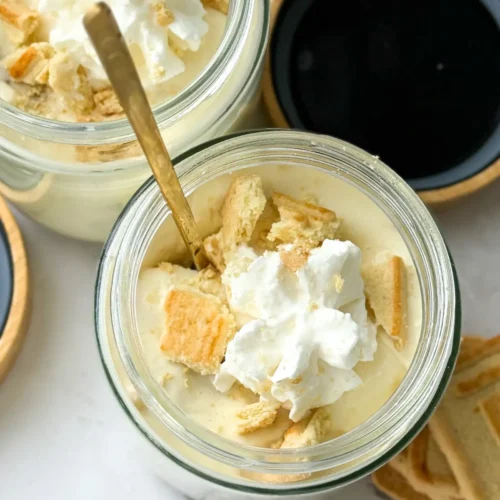 A birds-eye view of two Chessmen butter cookie high protein banana puddings in jars topped with whipped cream with gold spoos on each jar.