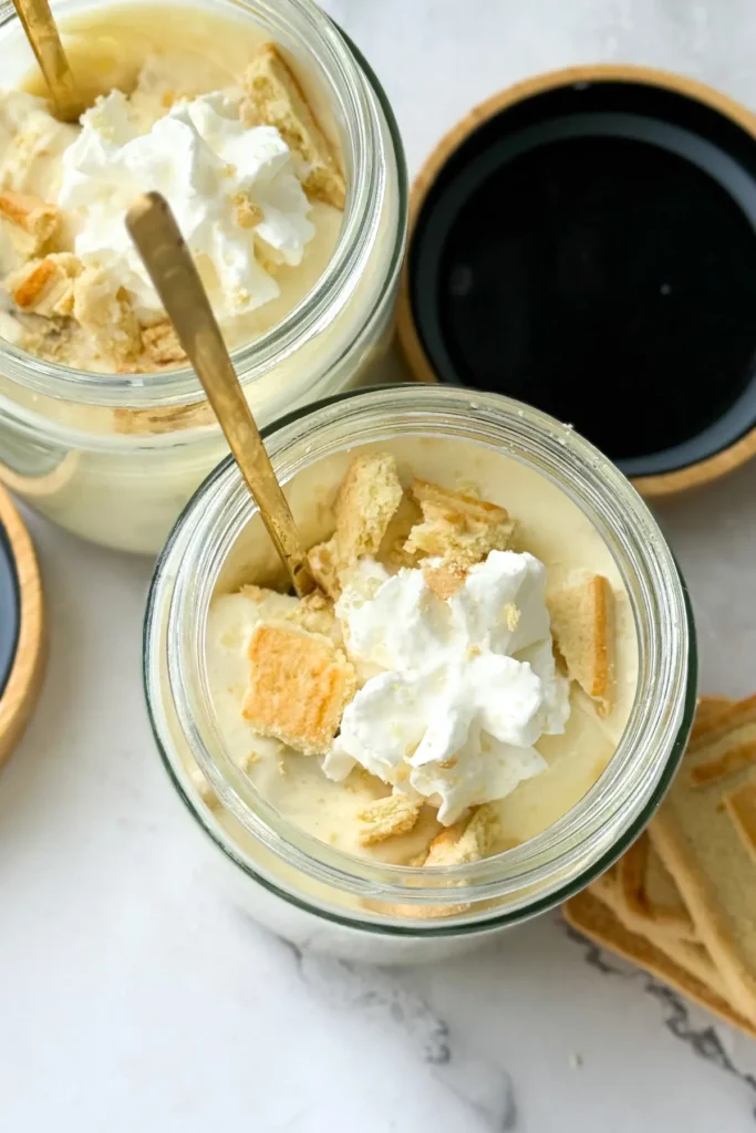 A birds-eye view of two Chessmen butter cookie high protein banana puddings in jars topped with whipped cream with gold spoos on each jar.