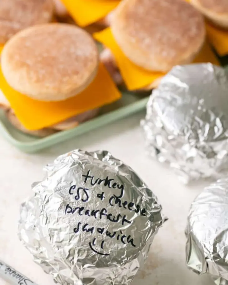 Make ahead breakfast sandwiches wrapped in foil with sharpie written on them with a pan of sandwiches in the background.