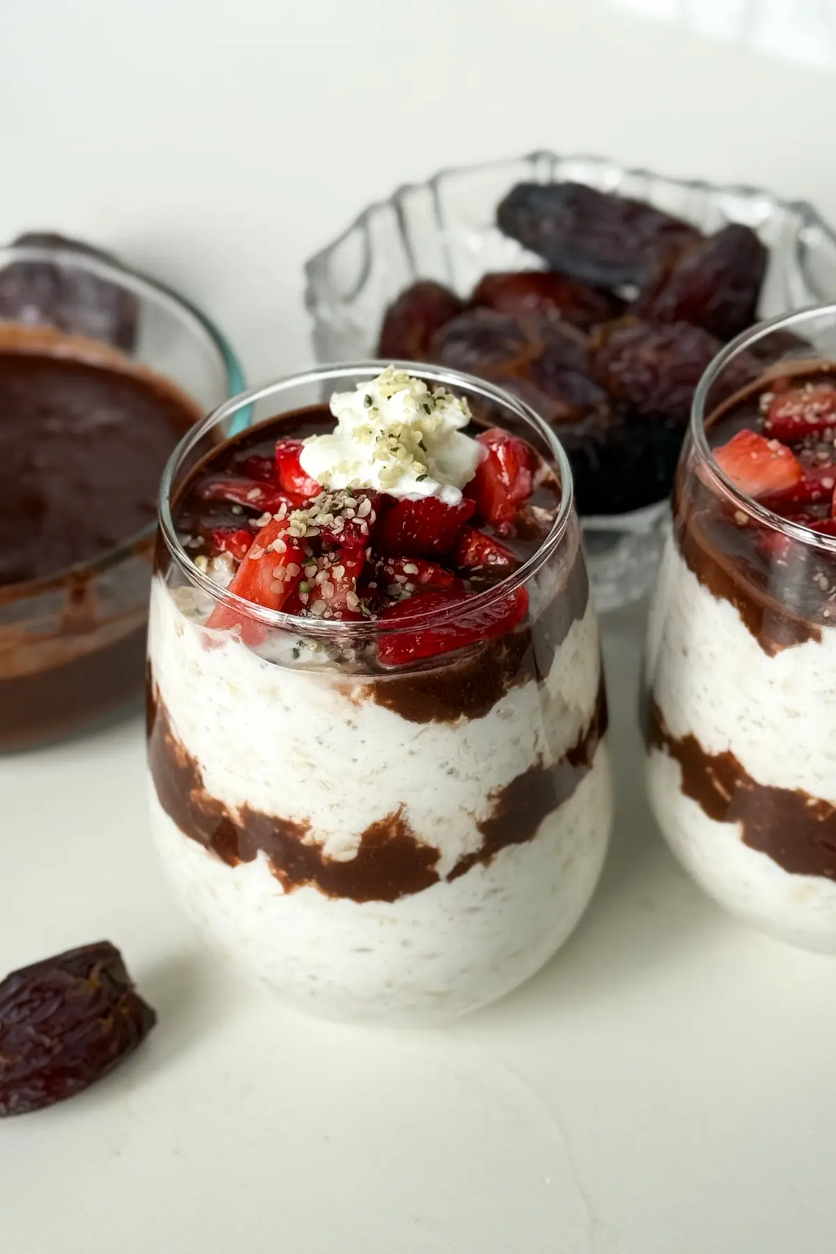 Two glasses of dark chocolate strawberry overnight oats with date sauce on a counter next to a bowl of date sauce.