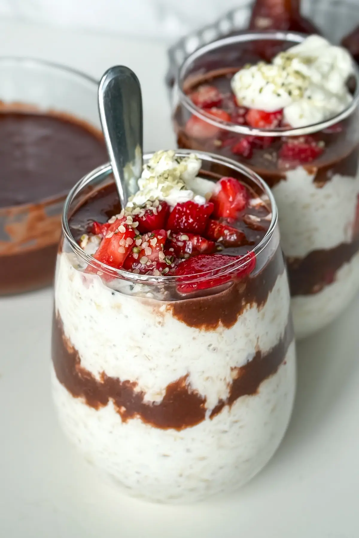 Two galsses of overnight oats layered with dark chocolate date sauce and diced strawberries next to a bowl of date sauce.