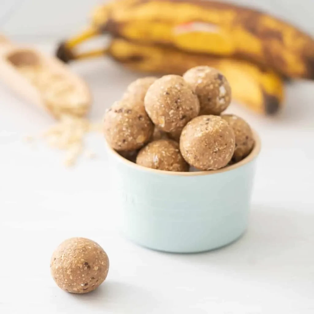 Banana bread balls stacked on top of one another in a small white bowl on a counter next to a bunch of bananas.