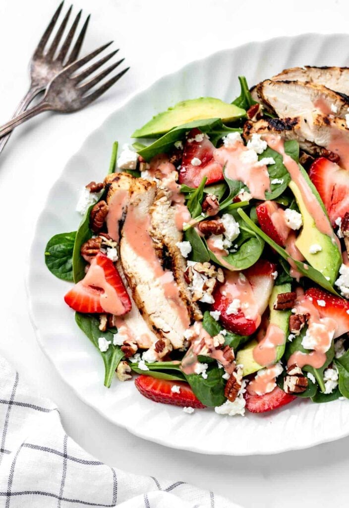 Grilled chicken over spinach with strawberries and chopped nuts in a white bowl on a table.