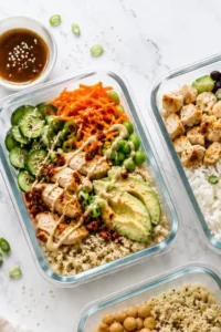 A close-up of a meal prep Asian chicken and rice bowl on a counter next to other meal prep containers and a bowl of sauce.