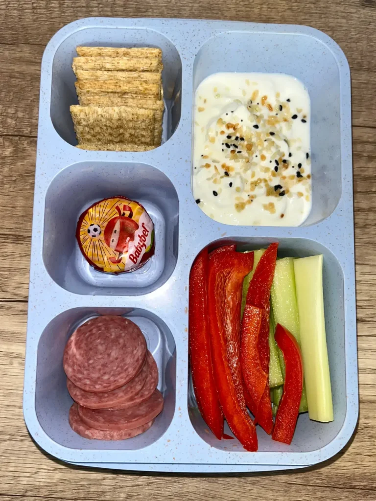 An adult lunchable consisting of salami slices, raw veggies, Greek yogurt dip, Baby Bel cheese and triscuit crackers.