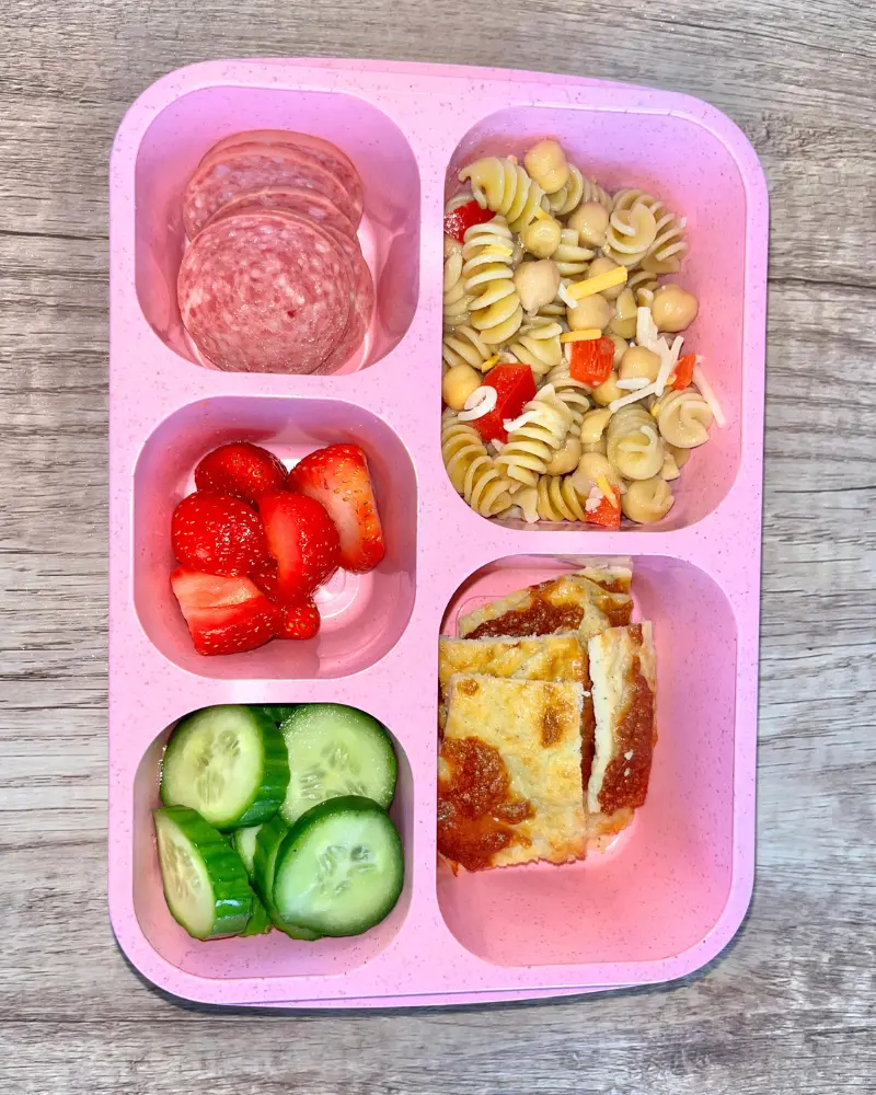 A lunchable featuring cucumber slices, strawberries, salami, pasta salad and cottage cheese flatbread.