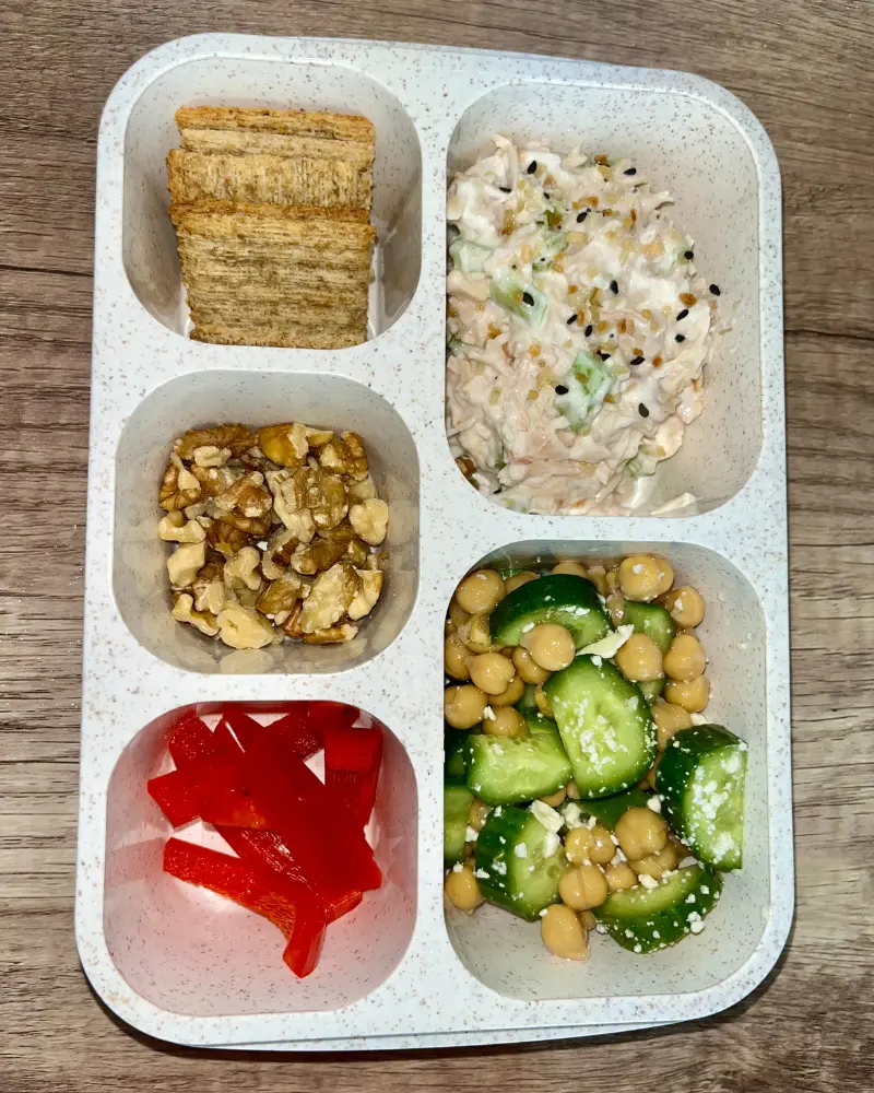 A 5 compartment Bento box with veggie slices, Greek yogurt chicken salad, Triscuits, chopped walnuts and a cucumber garbanzo bean salad.