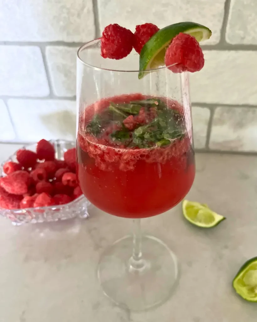 The basil berry spritzer kombucha mocktail in a wine glass on a counter next to a bowl of raspberries.