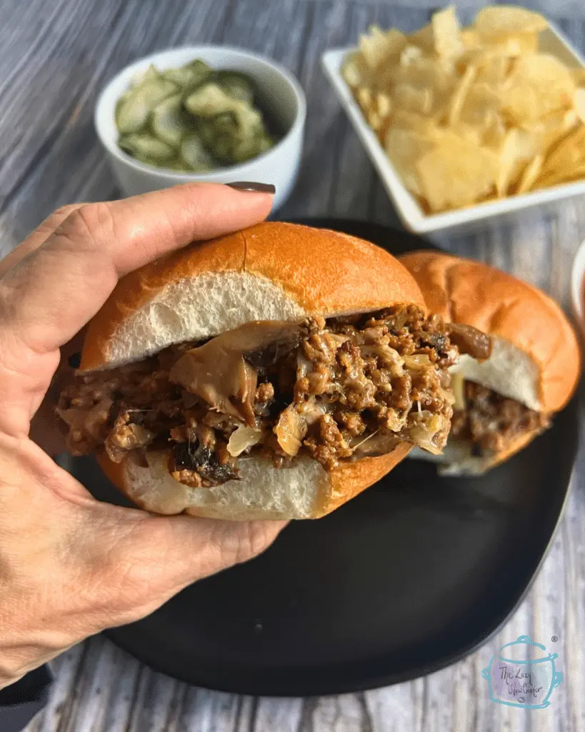 A cheesesteak sloppy joe on a bun in between someone's fingers with a plate in the background.