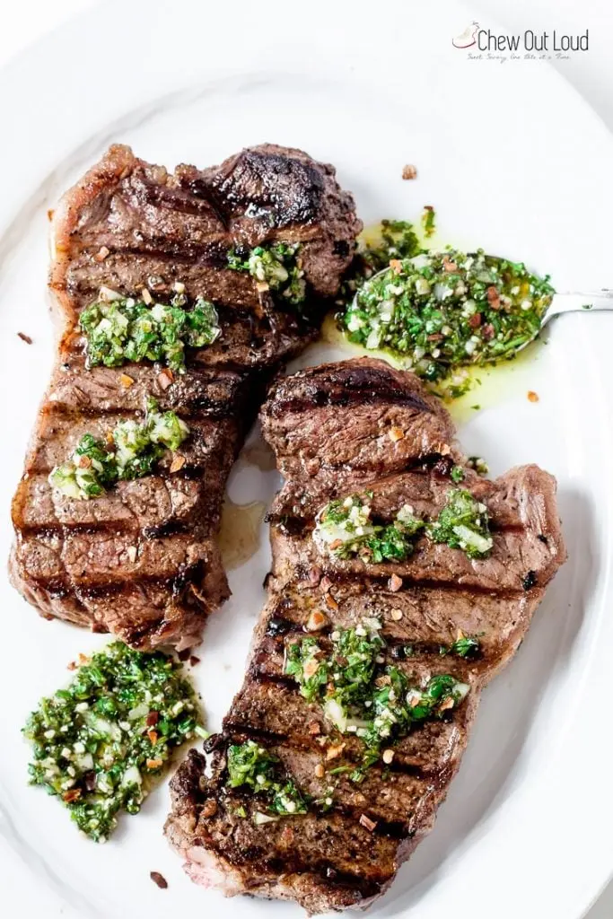 Two steaks on a white plate topped with chimichurri sauce.