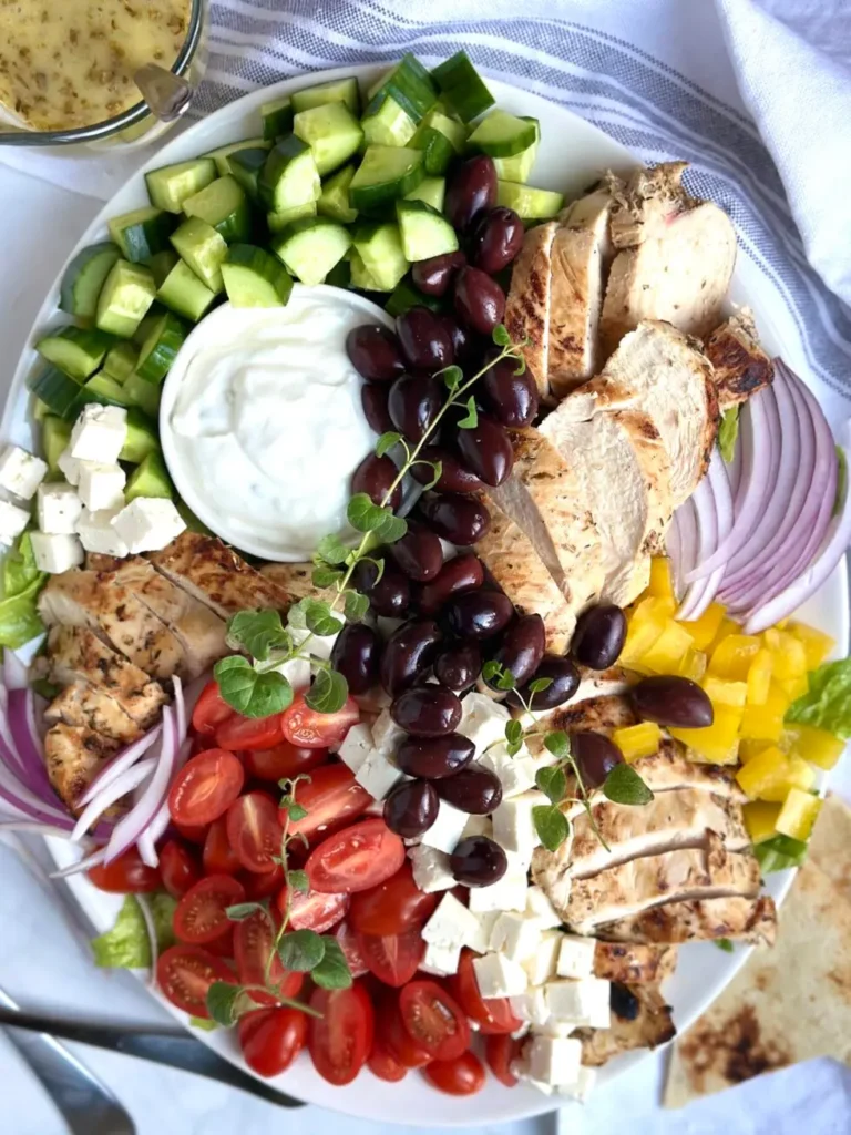 A Greek chicken salad with chopped veggies, olives, feta cheese, and a bowl of Tzatziki sauce on a plate.
