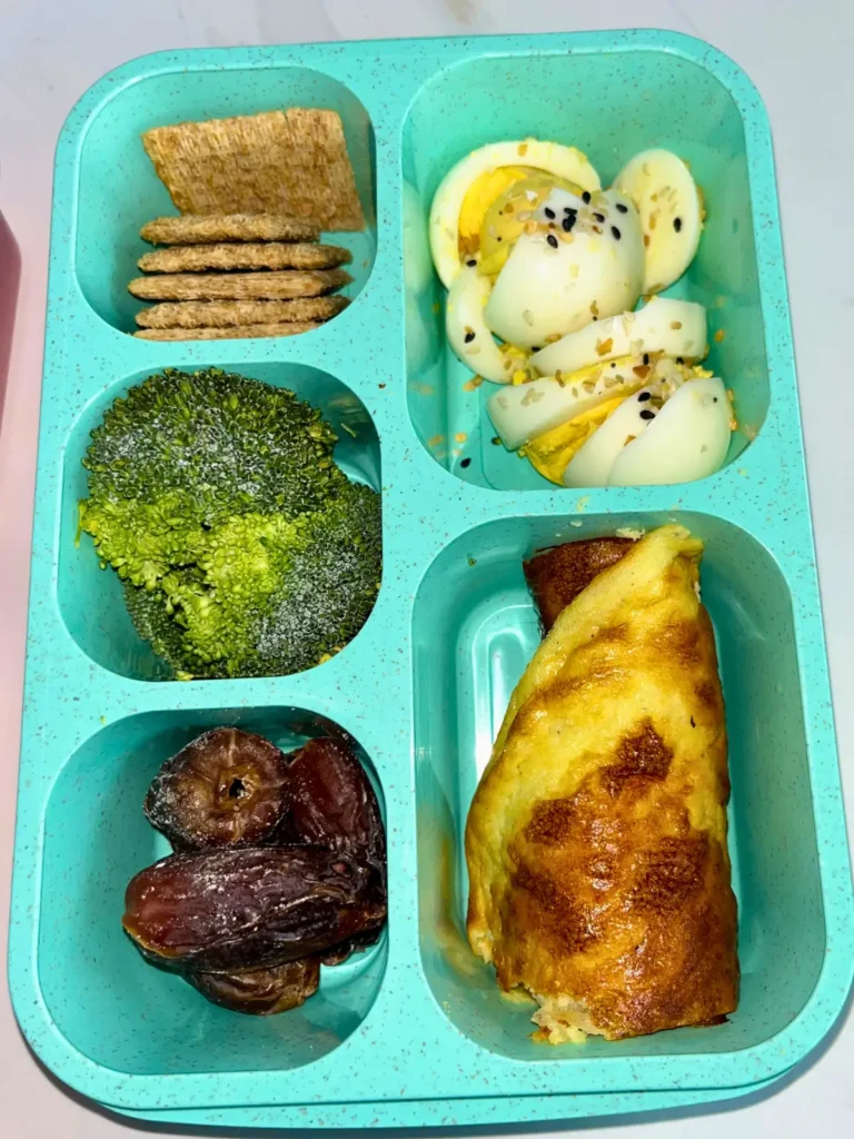 A high protein and fiber adult lunchable featuring hard boiled eggs, broccoli, pitted dates, cottage cheese flatbread and crackers.
