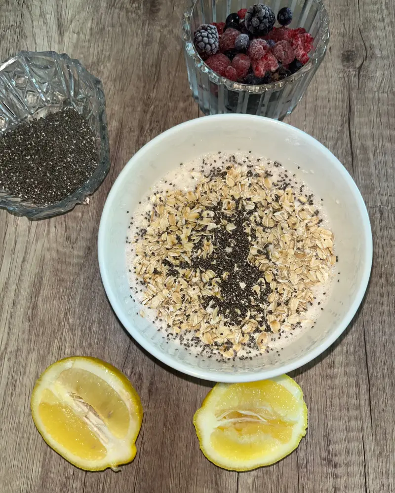 All the ingredients for the overnight oats mixture in a bowl on the counter next to lemon wedges.