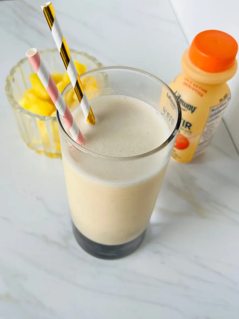 A peach pineapple kefir smoothie with two straws in it, and pineapple chunks and a bottle of kefir in the background.