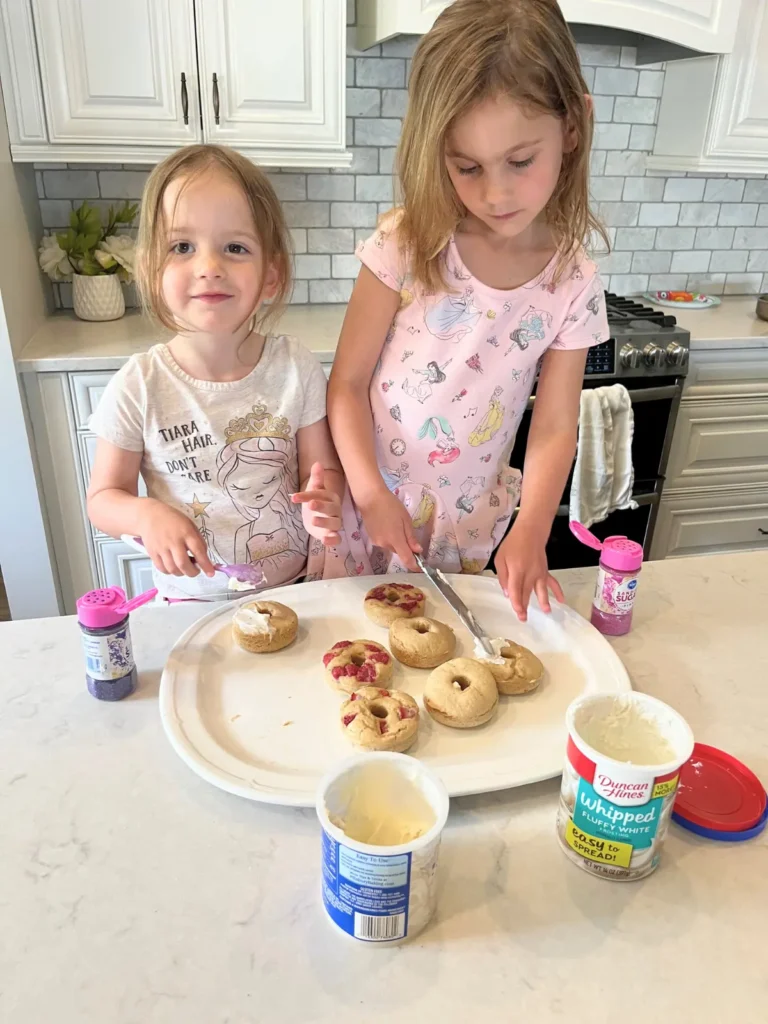 Two girls decorating protein donuts with frosting and sprinkles.