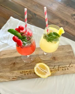 The basil berry spritzer and lemon ginger kombucha mocktail in wine glasses on a wood cutting board garnished with a lemon on a table.