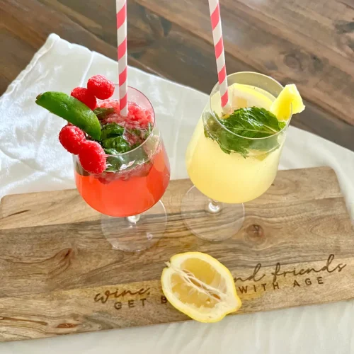 The basil berry spritzer and lemon ginger kombucha mocktail in wine glasses on a wood cutting board garnished with a lemon on a table.