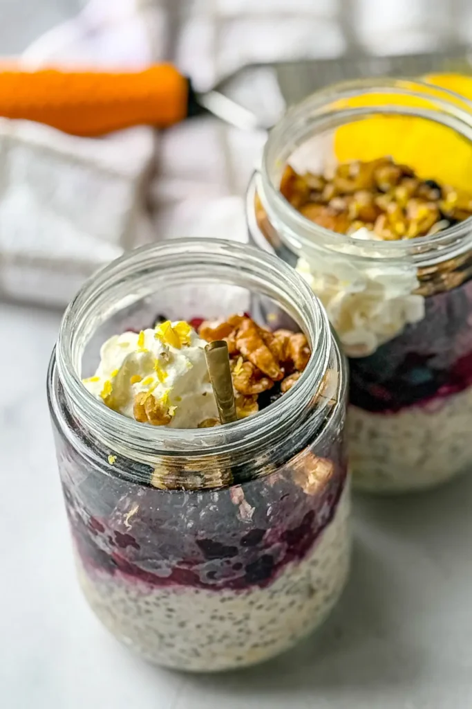 Two large mason jars with lemon berry kefir overnight oats topped with whipped cream and chopped walnuts on a counter next to a lemon zester. 