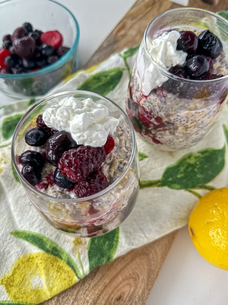 Two small glasses with lemon berry overnight oats made with kefir and protein powder on a wooden cutting board with a lemon decorated hand towel on it.