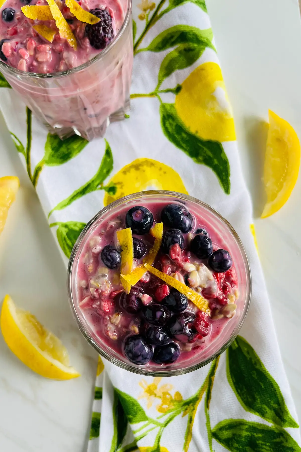 A birds-eye view of two glasses of fully assembled lemon berry overnight oats with kefir on a decorative lemon towel.