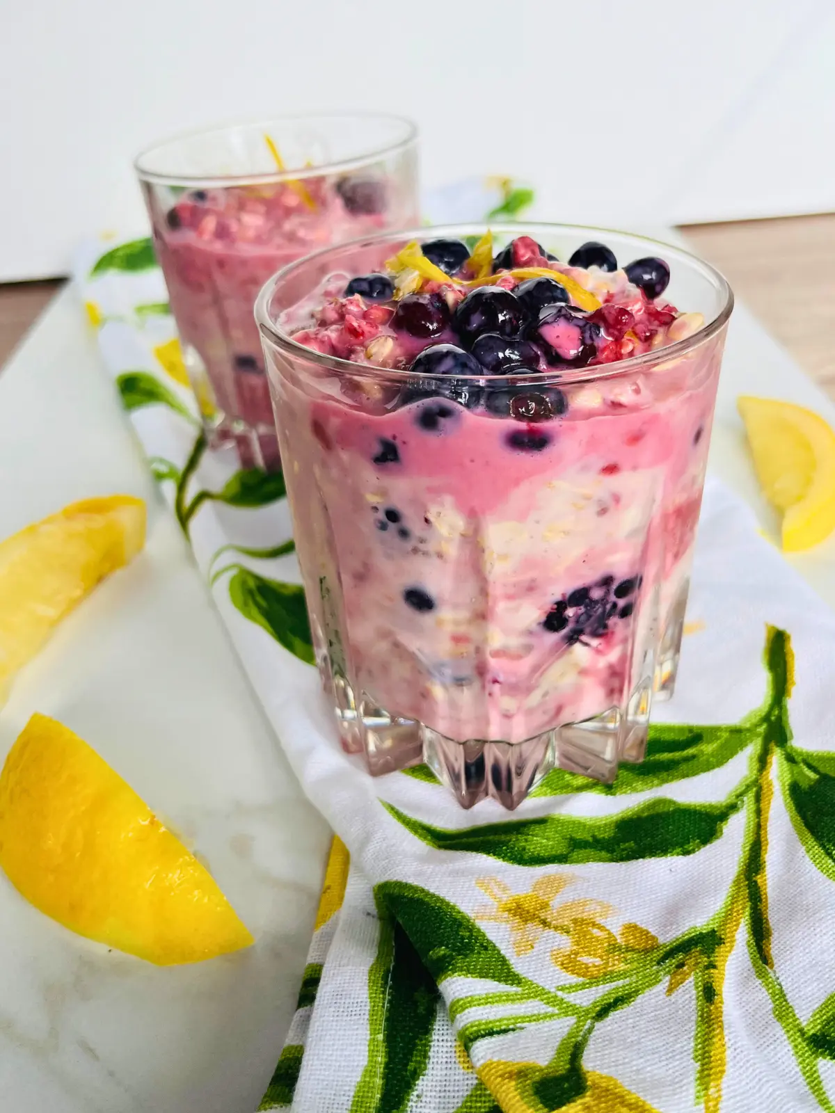 Two lemon berry overnight oats with kefir in glasses on a lemon towel on a kitchen counter.