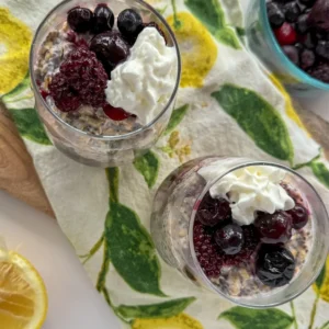 A birds-eye view of overnight oats in two glasses topped with mixed berries and whipped cream on a wooden cutting board with a lemon decorated hand towel on it.