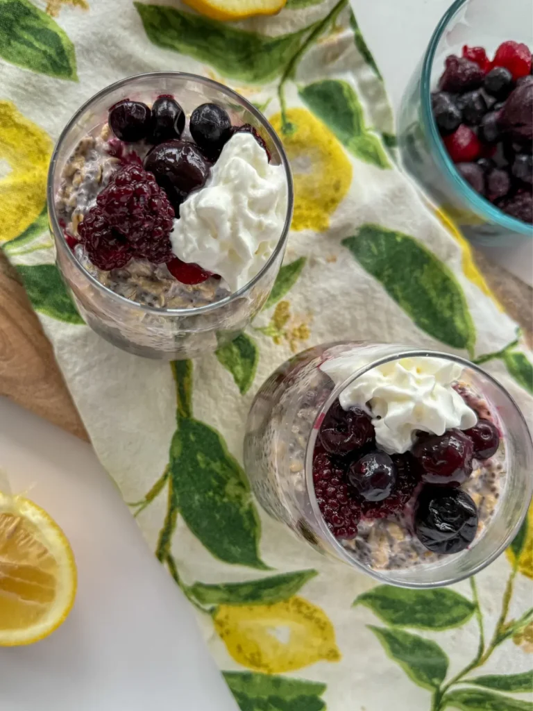 A birds-eye view of overnight oats in two glasses topped with mixed berries and whipped cream on a wooden cutting board with a lemon decorated hand towel on it.