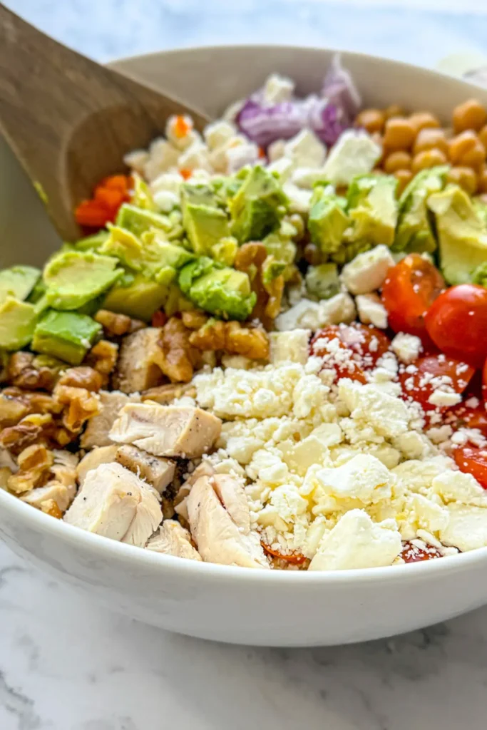 A close-up of a Mediterranean feta cheese and chicken salad with fresh vegetables in a large white bowl with a wooden spoon in it.