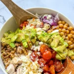 A birds-eye view of a chicken and veggie salad with feta cheese in a large bowl being mixed together with wooden spoons.