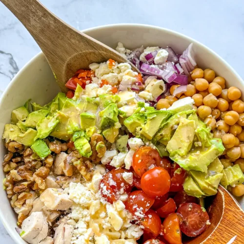 A birds-eye view of a chicken and veggie salad with feta cheese in a large bowl being mixed together with wooden spoons.