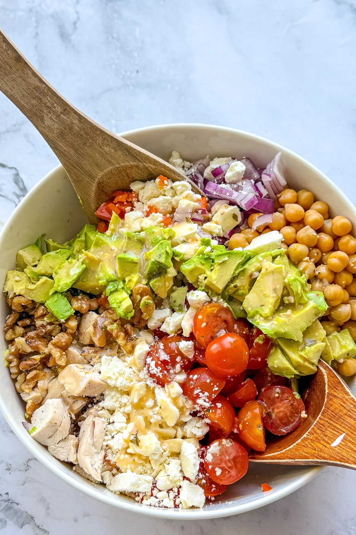 A birds-eye view of a chicken and veggie salad with feta cheese in a large bowl being mixed together with wooden spoons.