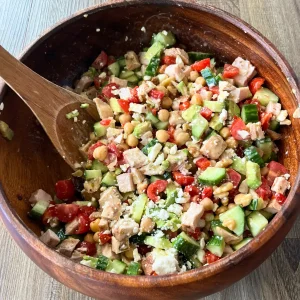 Mediterranean chicken, cucumber, and tomato salad in a wooden bowl with a wooden spoon in it.