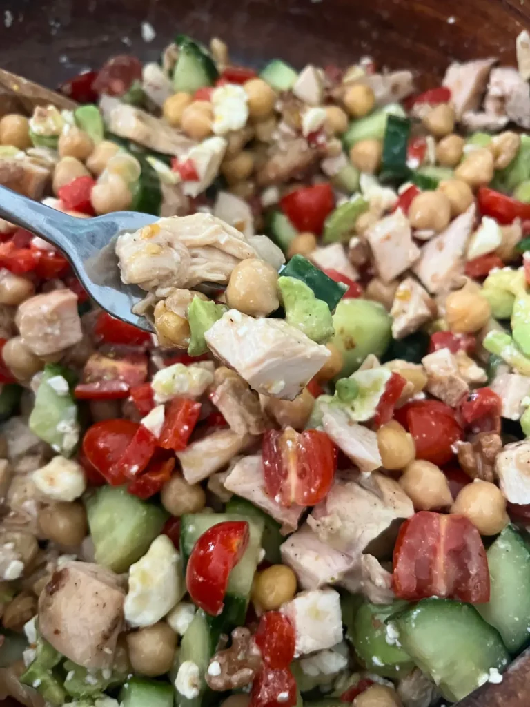 A close-up of a large forkful of Mediterranean chicken, cucumber and tomato salad with a large bowl of it in the background.