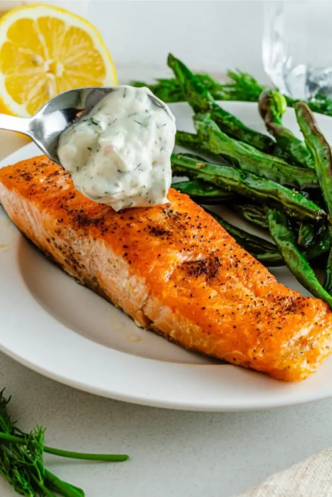 A salmon filet next to asparagus on a white plate with a spoonful of creamy dill sauce on top.