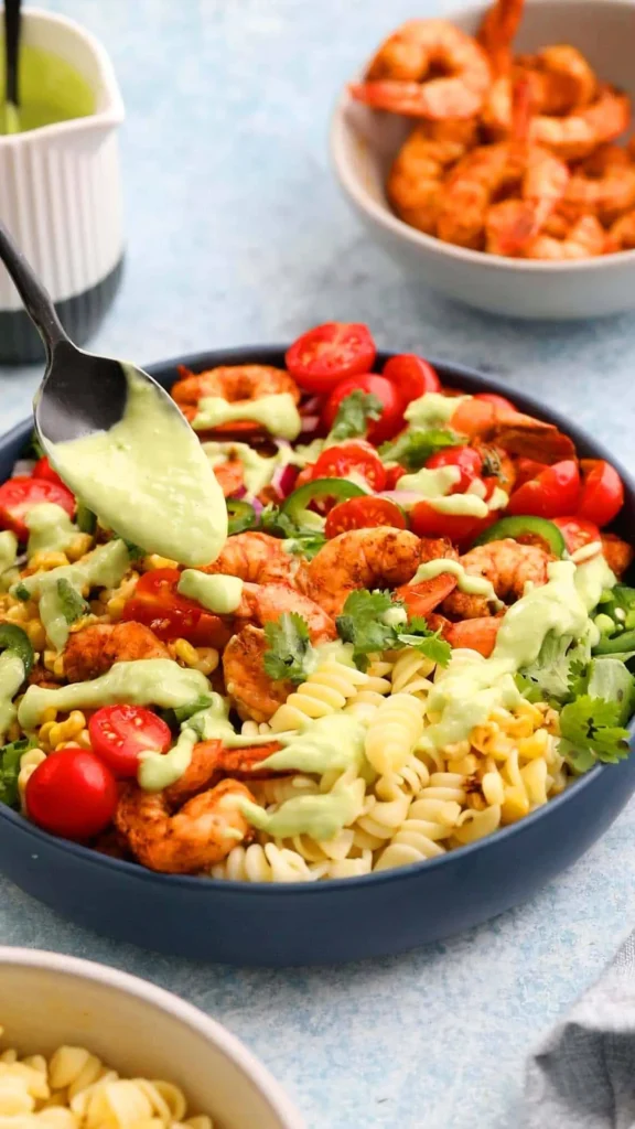 Shrimp pasta salad in a bowl with a spoon drizzling avocado dressing on it.